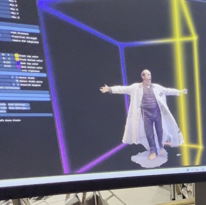A performer in a lab coat, arms outstretched, reconstructed as a volumetric point cloud inside a wireframe capture volume on screen